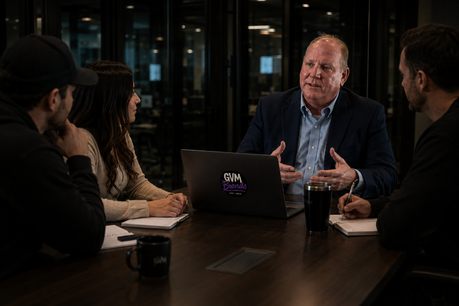 Four professionals sit around a wooden conference table in a dimly lit room, discussing with a laptop displaying a GVM Brands sticker at center.